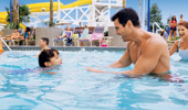 A girl, learning to swim, paddles toward her dad while her mom cheers.